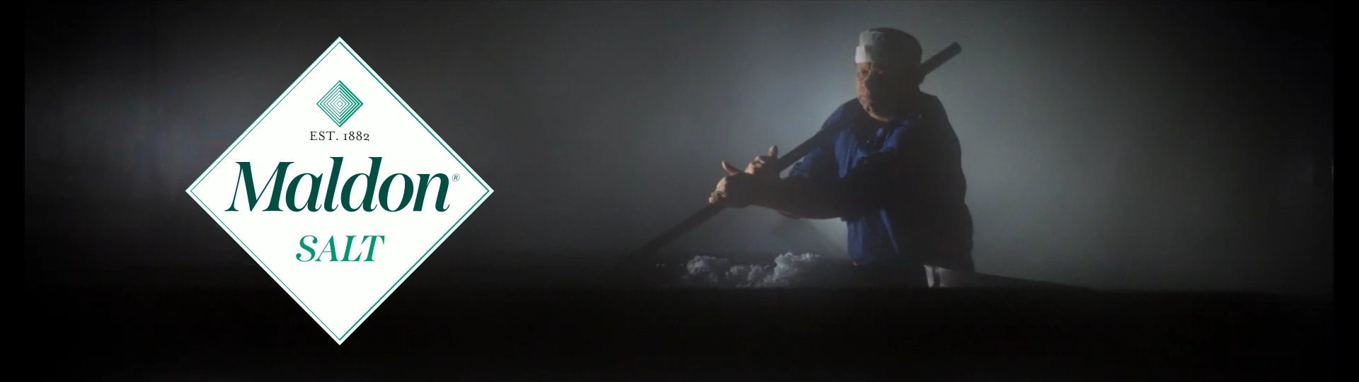 Maldon Sea Salt Kategorie-Teaser Ein Mann steht vor einem mystisch wirkenden, schwarz-weiß gehaltenen Hintergrund und schöpft mit einer Schaufel Salz aus einer flachen Salzpfanne. Links im Bild ist das Firmenlogo von Maldon Salt mit dem Schriftzug auf weißem Grund zu sehen.