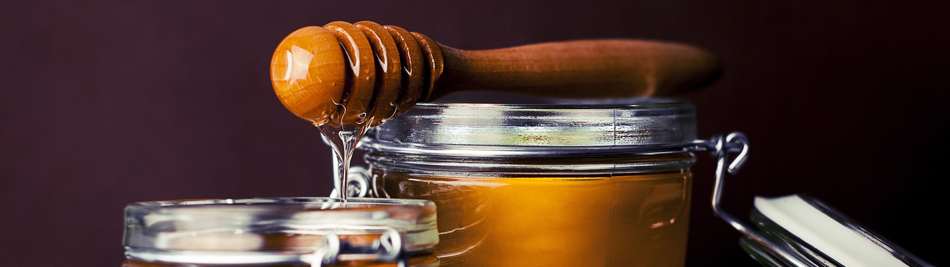 Glas mit goldgelbem Honig, kunstvoll angerichtet mit einem Holzlöffel, tropfend über den Rand, vor einem rustikalen Hintergrund – ideal für Süßspeisen und Frühstück.