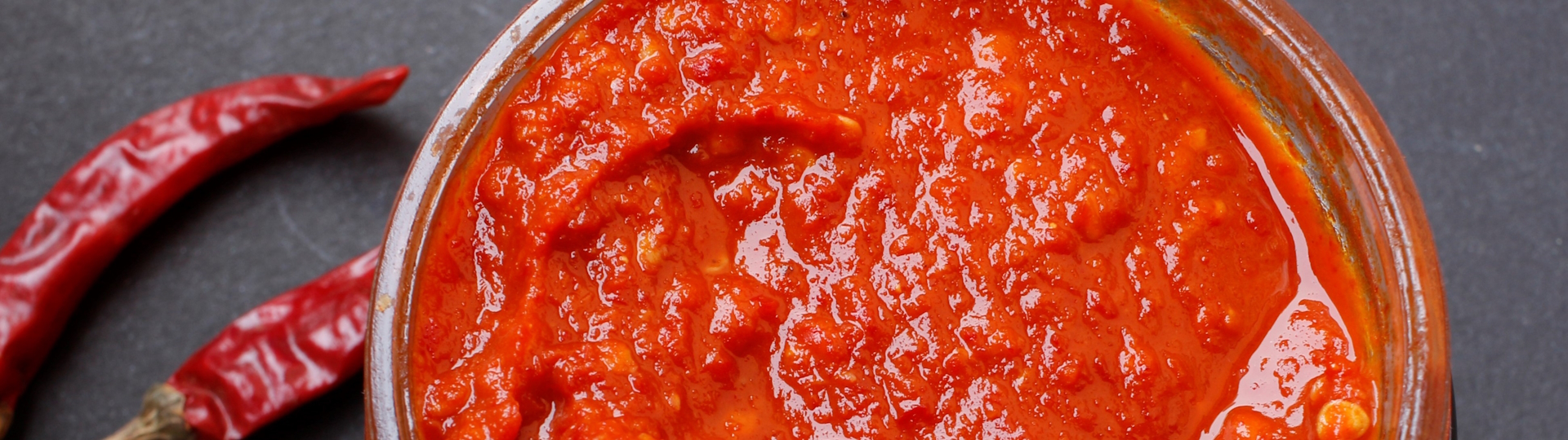 Aromatische Chilipaste – Würzige Schärfe aus hochwertigen Chilischoten Glas Chilipaste mit intensiver roter Farbe, präsentiert vor einem rustikalen Hintergrund, reich an frischen Chilischoten und ideal für scharfe Gerichte.